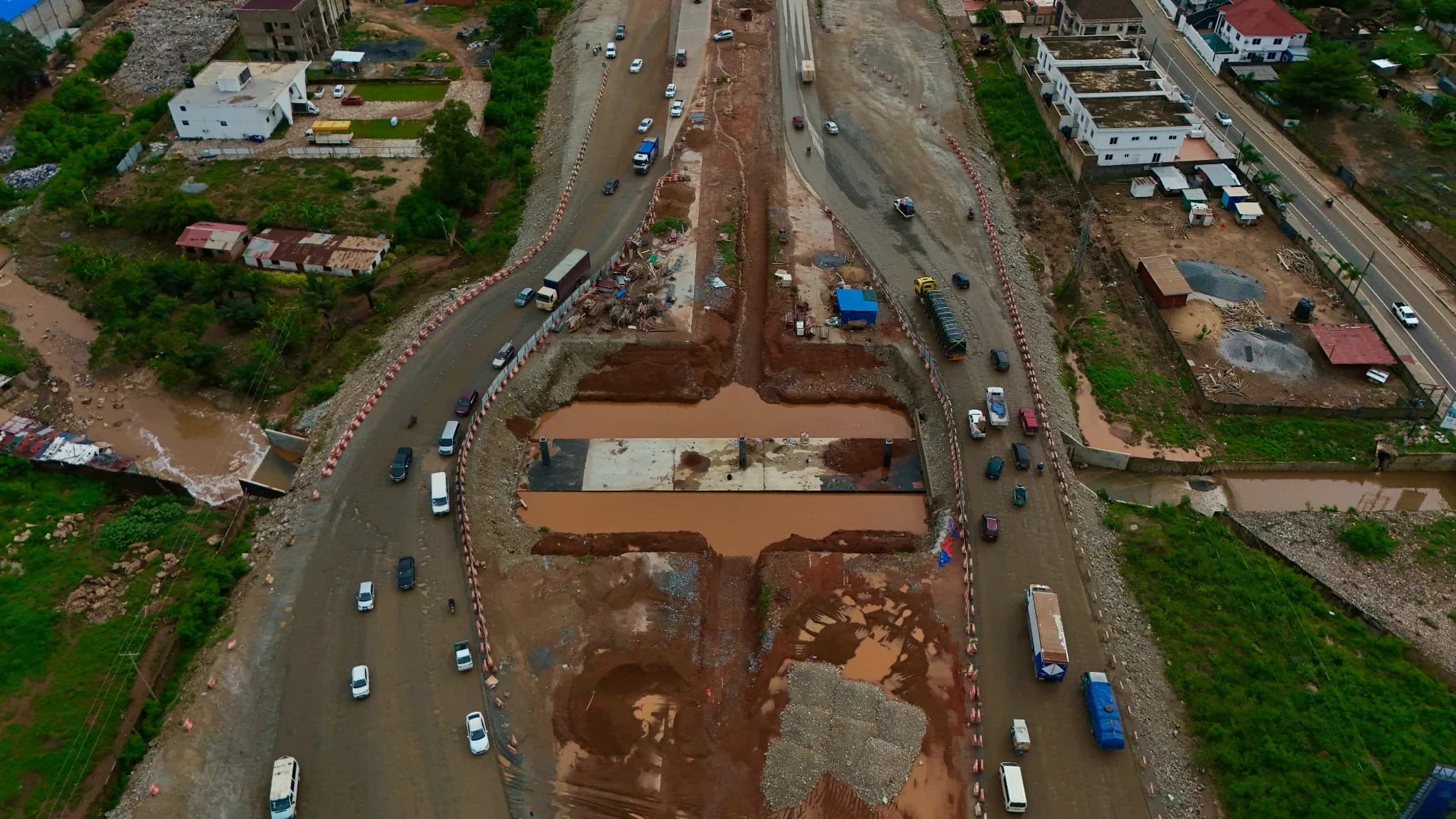 Aerial view of a road underpass under construction at a junction, with a concrete base slab in the excavation and traffic diverted around it on temporary lanes