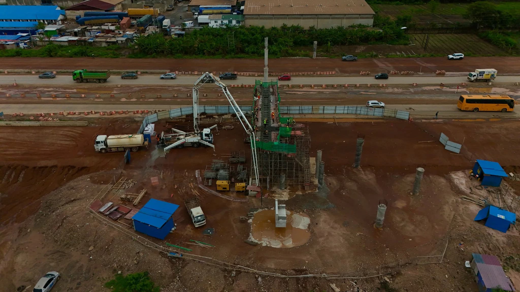 Aerial view of a bridge pier being concreted by a truck-mounted concrete pump beside the corridor, with traffic passing on the adjacent carriageway