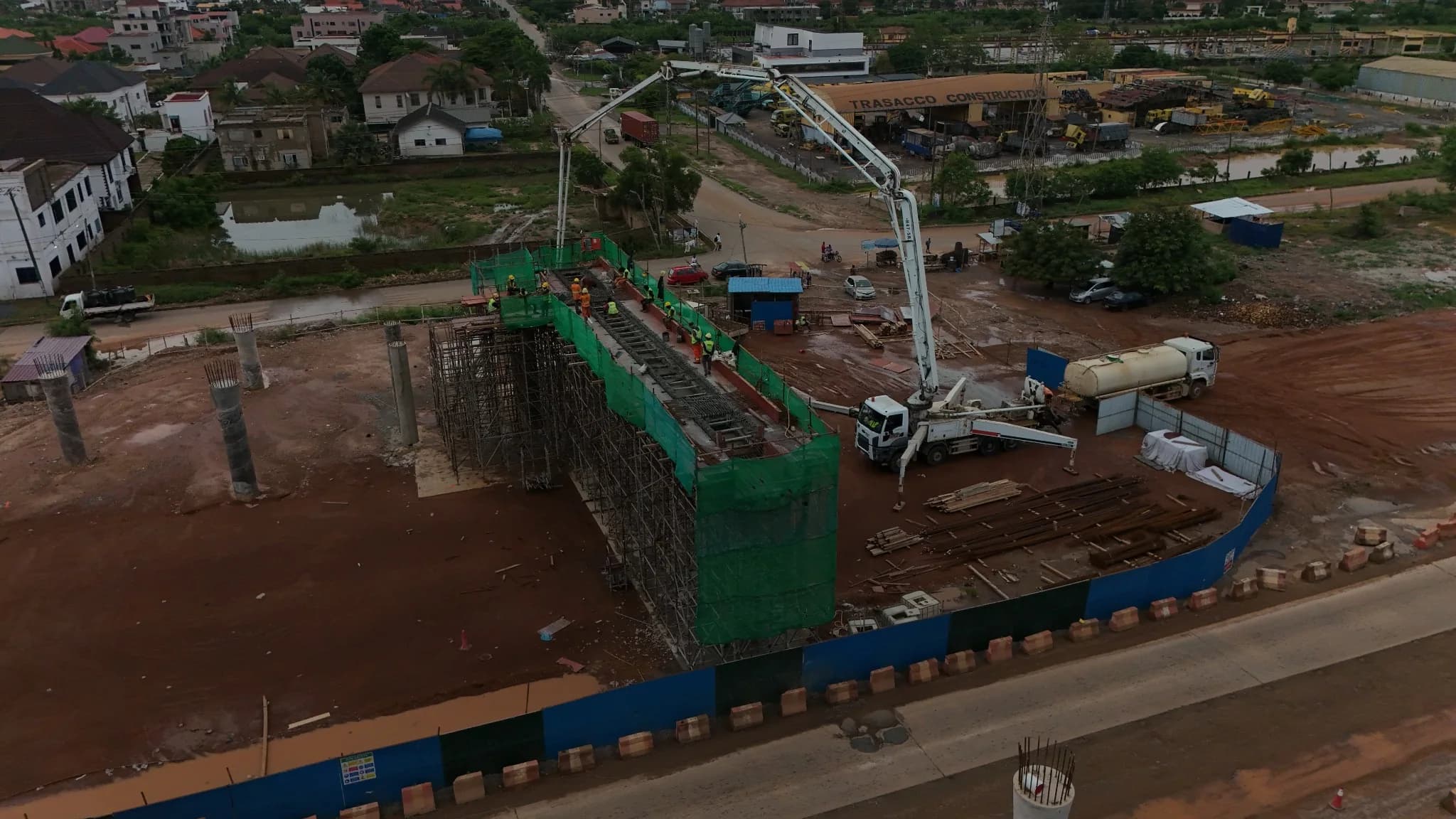 Aerial view of an overpass deck under construction, wrapped in green safety netting, with a concrete pump delivering to workers on the deck