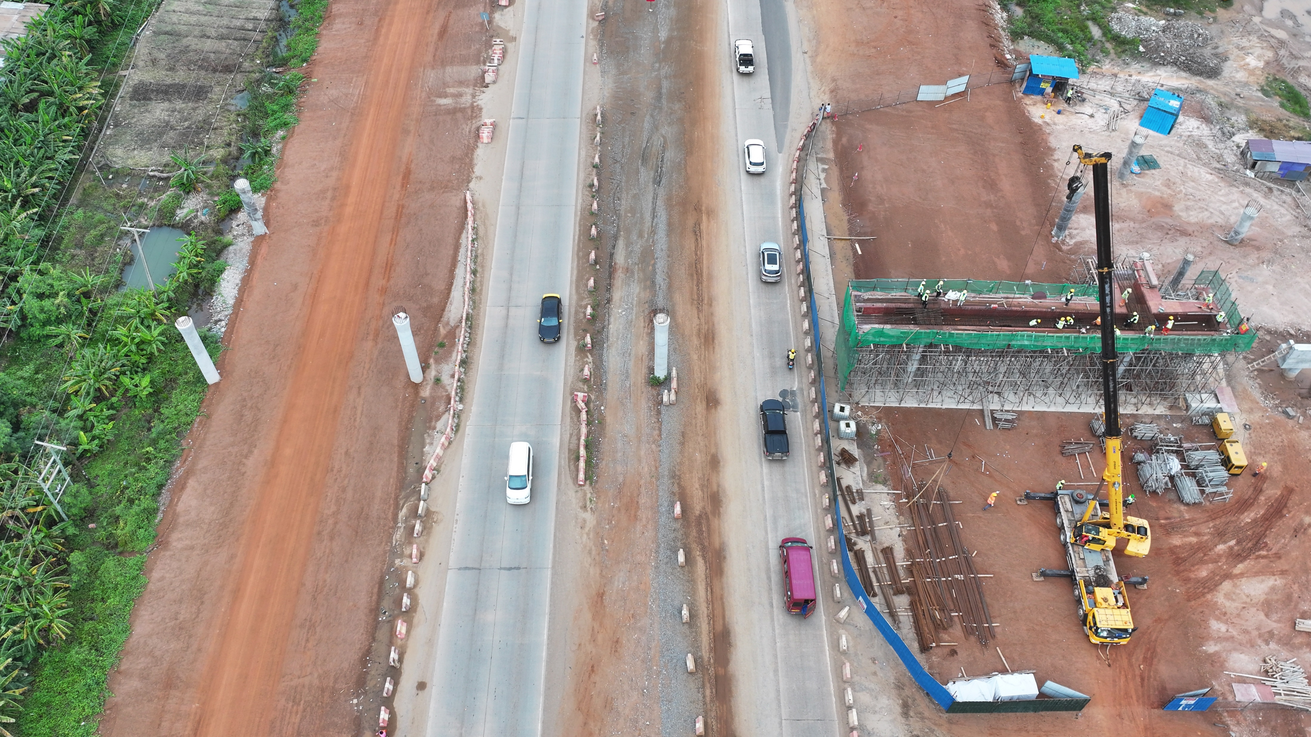 Aerial view of overpass pier and deck construction beside a live road, with a mobile crane and reinforcement cages