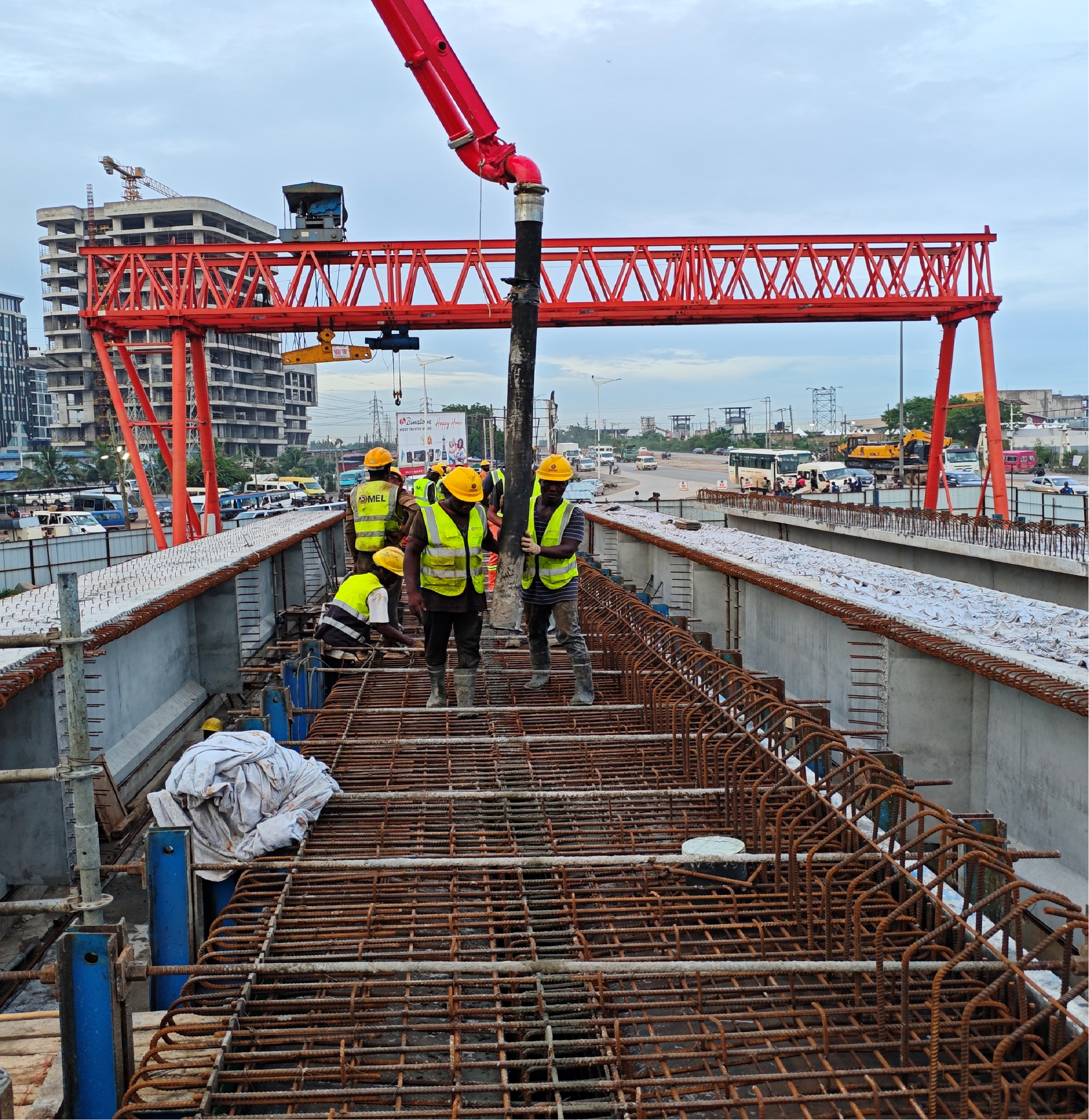 Bridge deck concreting beneath a red launching gantry, with a concrete pump and work crew on the deck