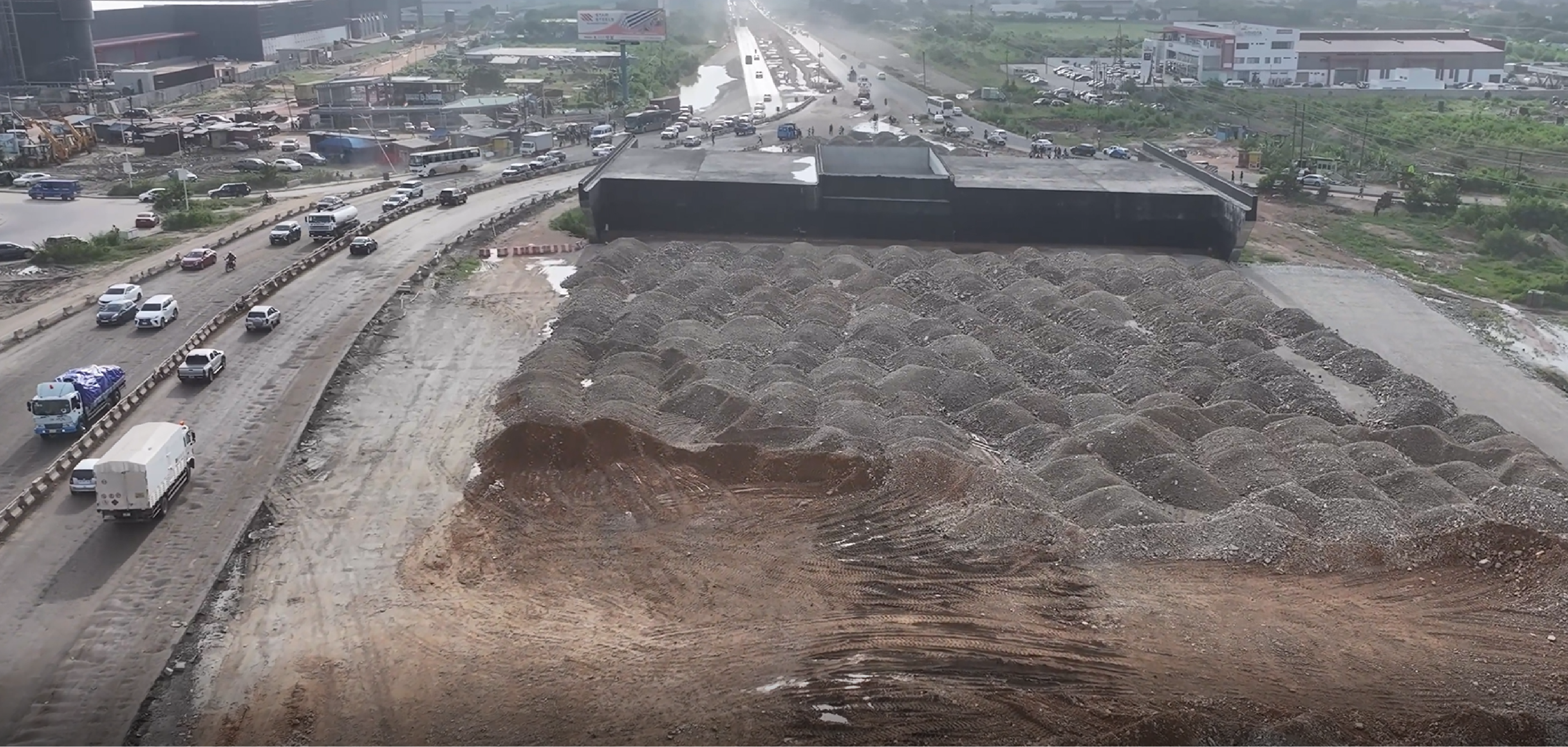 Aerial view of stockpiled rock fill and earthworks beside the corridor, with an underpass structure and traffic