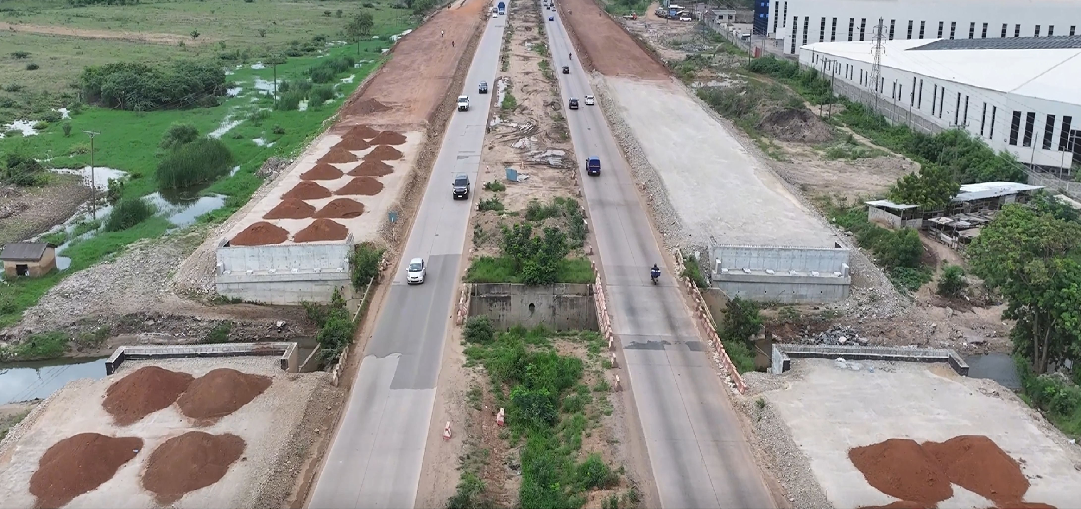 Aerial view of box-culvert construction and earth stockpiles alongside an existing road carrying traffic