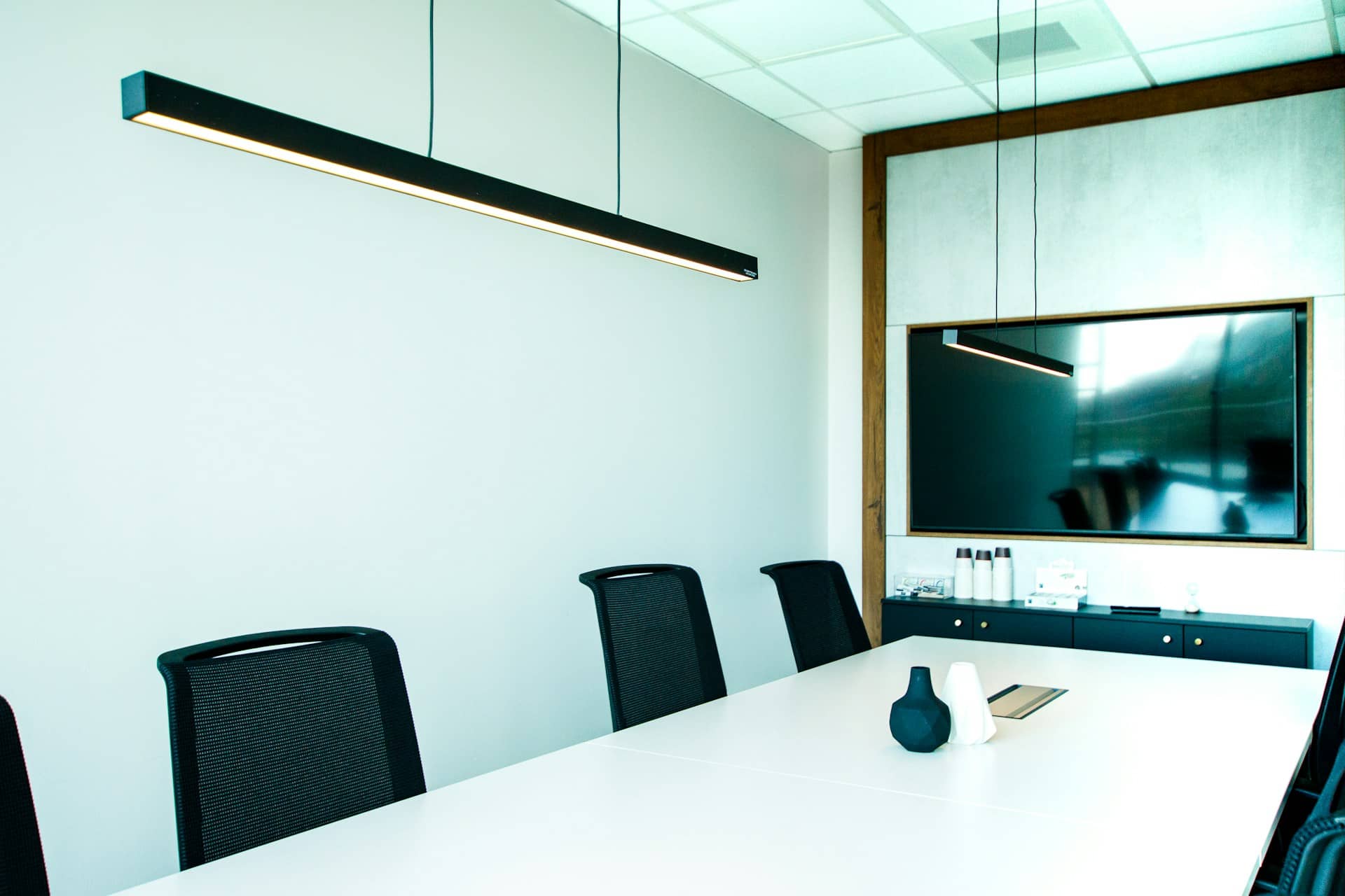 A bright, empty meeting room with a long white table, black mesh chairs, a pendant light and a wall-mounted screen