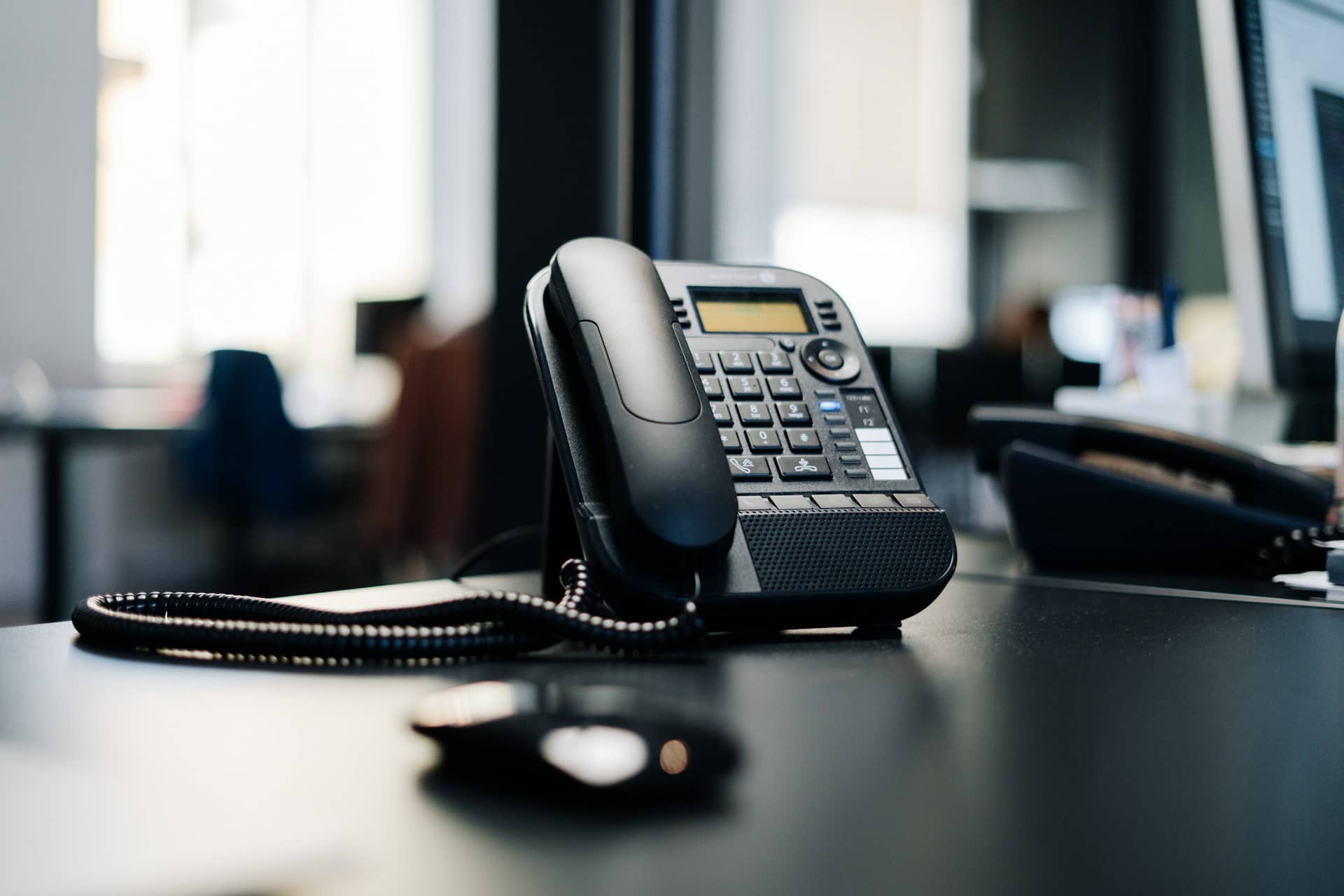 A black desk telephone with a coiled cord on an office desk, with a computer monitor blurred behind it