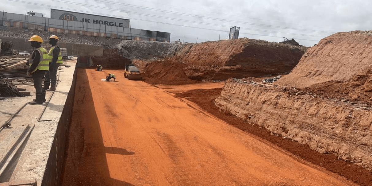 Workers in hi-vis vests beside a concrete wall as a small roller compacts a red-earth base in an excavated section of the corridor